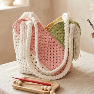 May include: A multi-coloured crochet tote bag featuring diagonal panels of cream, soft pink, and sage green yarn. The bag has thick, white braided rope handles with long fringe tassels at the attachment points. A red crochet hook rests on a wooden tray.
