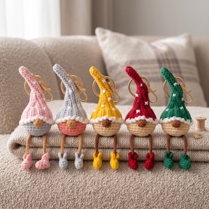 May include: Five crochet gnome keychains with gold-tone metal lobster clasps. Each gnome features a tall, pointed hat in pink, gray, yellow, red, or green with white star accents, a round nose, and dangling legs with matching colored boots.