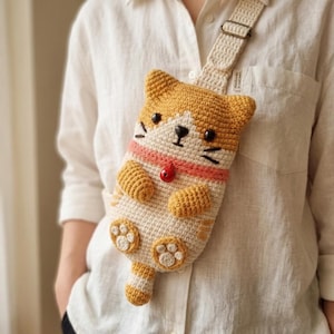 May include: A crochet crossbody bag shaped like a ginger and white cat with black eyes, a small nose, whiskers, and a pink collar featuring a red bell. The bag has a long adjustable strap, two small front paws, and a tail extending from the bottom.
