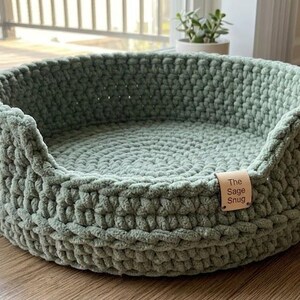 May include: A sage green, round pet bed with a thick, textured crochet design. The bed has a low profile with a slightly raised edge. A small, rectangular tag with the text "The Sage Snug" is attached to the front.