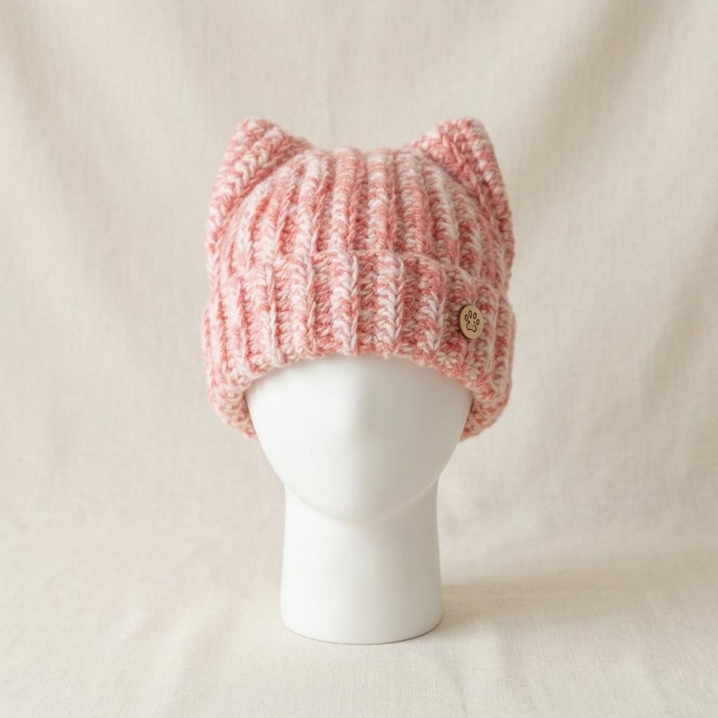 May include: A chunky knit beanie in a variegated pink and white yarn featuring two pointed cat-ear shapes at the top. The hat has a ribbed texture and a small, round, light-colored wooden button on the side embossed with a paw print design.