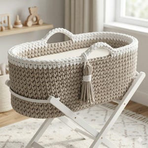 May include: An oval-shaped baby bassinet featuring a beige crocheted exterior with a thick white crocheted rim and two matching handles. A beige tassel hangs from one handle. The bassinet rests on a white wooden X-frame stand with a flat mattress.