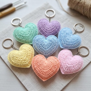 Può includere: Una collezione di sette portachiavi a forma di cuore realizzati in filato lavorato all'uncinetto in colori pastello, tra cui verde menta, lavanda, azzurro, giallo pallido, pesca e rosa tenue, ciascuno fissato a un anello in metallo argentato.