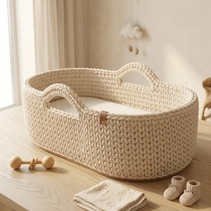 Peut inclure: Un couffin pour bébé tricoté de couleur crème, de forme ovale, avec des poignées intégrées et une languette en cuir marron. Le couffin contient un matelas blanc lisse et est accompagné d'un hochet en bois, d'un tissu plié et de deux chaussons.