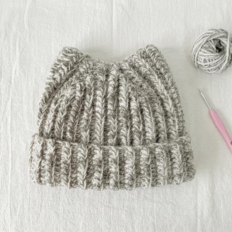 May include: A grey and white variegated yarn beanie with a ribbed knit texture and two pointed cat-ear style peaks at the top. The hat is displayed alongside a matching ball of yarn and a pink crochet hook, showcasing the handmade construction.