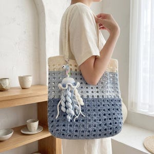 May include: A crochet tote bag featuring a horizontal ombre color gradient transitioning from cream at the top to light blue and dark blue at the base. The bag has an open-weave pattern and includes a hanging jellyfish-shaped crochet charm.