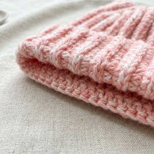 May include: A soft, hand-crocheted beanie hat featuring a ribbed texture and a variegated color pattern of light pink and white yarn. The knit construction displays a consistent, repeating stitch pattern throughout the folded brim and crown.
