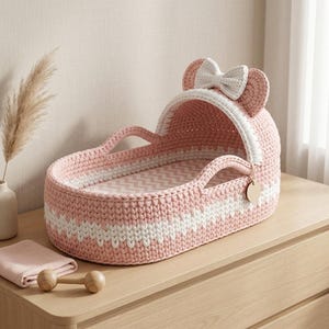 May include: A pink and white crocheted bassinet featuring a hood with two rounded ears and a central white bow. The interior base has a chevron pattern in pink and white. A small circular wooden tag hangs from the side of the hood structure.