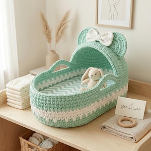 May include: A mint green crocheted bassinet with a white bow on the hood, featuring a chevron patterned mattress and a small beige crocheted rabbit toy inside. A white card with the text Stitchora Crochet is placed next to a wooden ring.