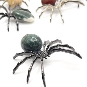 May include: Several decorative spider figurines with gemstone bodies and dark metal legs are displayed. The gemstones are various colors, including green, red, yellow, and purple. The spiders are positioned on a white surface.