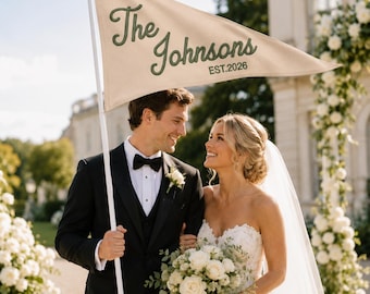 May include: A triangular beige pennant flag attached to a white pole, featuring the dark green embroidered text The Johnsons and EST.2026. The flag is displayed outdoors, showcasing a clean fabric texture and bold, cursive lettering for the family name.