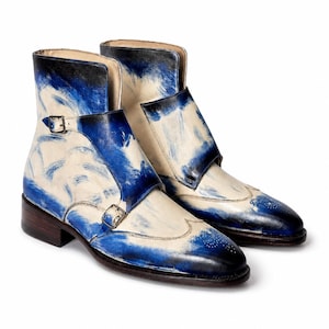 Op de afbeelding: Een paar enkellaarzen met een dubbele gespsluiting en zilverkleurige gespen. De laarzen hebben een crèmekleurige basis met een handgeschilderd blauw penseelstreekpatroon, een wingtip neus met perforaties en een donkerbruine hak.