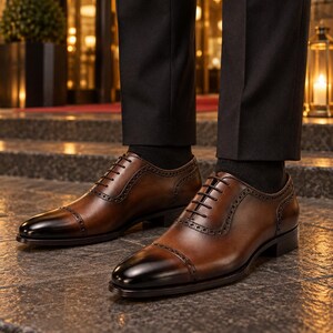 Men’s polished brown leather Oxford shoes with dark cap toes and brogue trim worn with black trousers outside an elegant hotel.