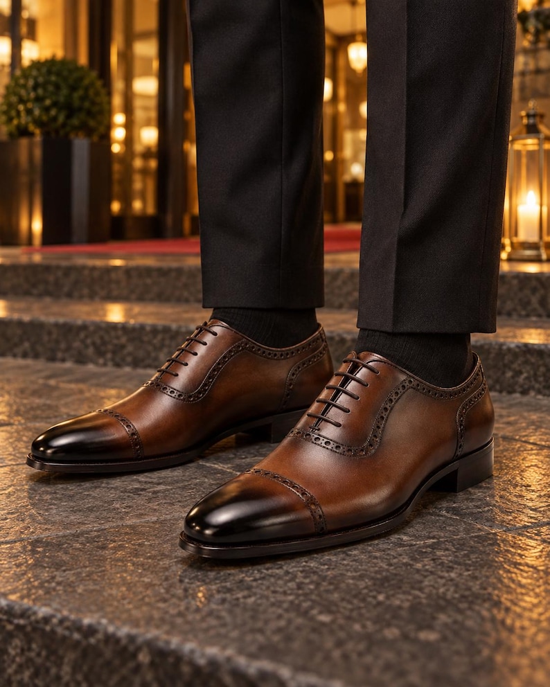 Men’s polished brown leather Oxford shoes with dark cap toes and brogue trim worn with black trousers outside an elegant hotel.