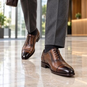 Men’s brown leather Oxford dress shoes with dark burnished cap toes worn with charcoal trousers while walking through a modern office lobby.