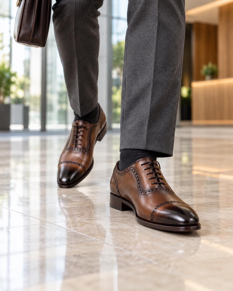 Men’s brown leather Oxford dress shoes with dark burnished cap toes worn with charcoal trousers while walking through a modern office lobby.