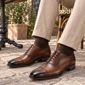 Men’s brown cap-toe Oxford shoes styled with beige chinos and dark socks while seated at a relaxed outdoor café.