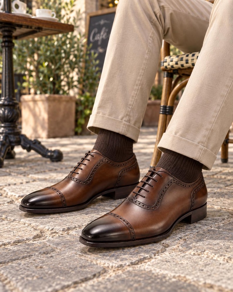 Men’s brown cap-toe Oxford shoes styled with beige chinos and dark socks while seated at a relaxed outdoor café.
