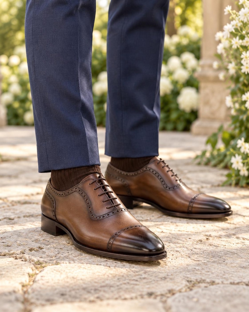Men’s brown leather cap-toe Oxford shoes worn with navy suit trousers at an outdoor garden wedding, featuring dark burnished toes and brogue detailing.