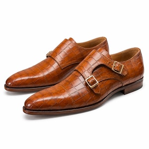 May include: A pair of tan double monk strap shoes featuring a textured crocodile-embossed pattern. The shoes have a pointed toe, a low stacked heel, and two gold-toned metal buckles on the side of each shoe, securing the leather strap across the top.