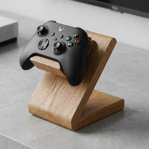 Op de afbeelding: Een zwarte Xbox-controller rust op een houten standaard. De controller heeft gekleurde knoppen en een D-pad. De standaard is gemaakt van lichtgekleurd hout met een Z-vormig ontwerp, wat een stijlvolle weergave biedt voor gaming-accessoires.