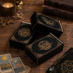 May include: Three rectangular black wooden boxes featuring gold-coloured engravings. One box displays a moth, another a circular emblem with a central eye, and the third a pentagram design. Each box has decorative corner accents and a dark finish.