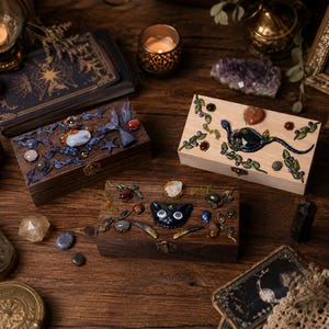 May include: Three rectangular wooden boxes with hinged lids and metal latches. One dark wood box features a mermaid tail and stones, a light wood box displays a snake and heart, and a dark wood box shows a black cat face with various stones.
