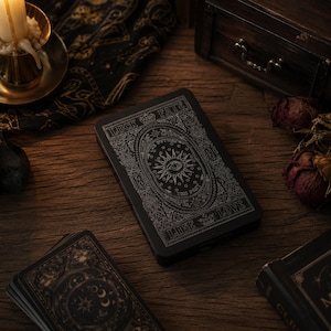May include: A rectangular black deck box featuring intricate silver line art of a central eye, sun rays, and stars. The text LUCK, FATE, LIFE, and LOVE is printed on the borders. The box is surrounded by tarot cards, a candle, and dried roses.