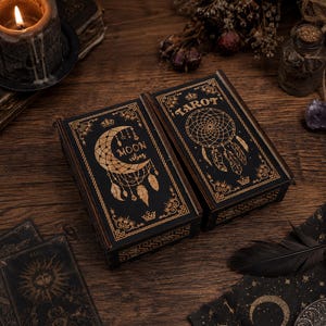 May include: Two rectangular wooden boxes with black lids featuring gold-coloured engravings. One lid displays a crescent moon and the text "MOON vibes," while the other lid shows a dreamcatcher design and the word "TAROT" with crown accents.