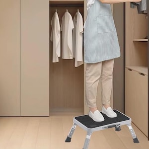 May include: A foldable, silver and black step stool with adjustable legs. The step stool has a textured black surface and is designed to provide a stable platform for reaching higher places. It is shown in a closet setting.