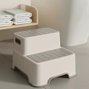 May include: A two-step white and grey step stool. The top surfaces have a textured, non-slip design. A small handle is on the side. The stool is designed for use in a bathroom or similar setting.
