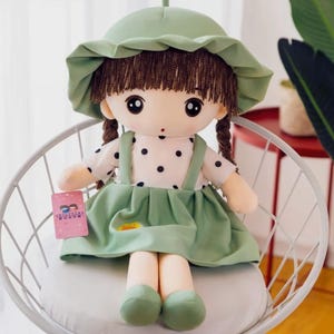 Pode incluir: Uma boneca de pelúcia com cabelo castanho trançado, usando um chapéu verde, um vestido branco com bolinhas e uma saia verde. A boneca está sentada em uma cadeira branca. Uma etiqueta rosa está presa ao vestido da boneca.