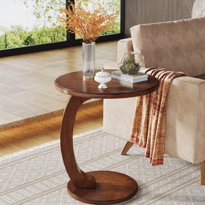 Peut inclure: Une table d'appoint ronde en bois avec une base en forme de C. Sur le plateau se trouvent un grand vase en verre cannelé transparent contenant du feuillage orange séché, un petit pot décoratif blanc à couvercle et deux livres superposés.