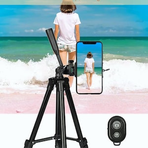 May include: Black tripod with a smartphone holder and a remote control. The tripod is set up on a beach, with a person walking in the background. The smartphone displays the same person on the beach.