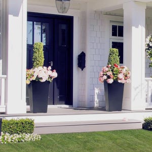 Puede incluir: Dos jardineras rectangulares negras llenas de flores rosas y blancas y topiarios verdes flanquean una puerta principal azul marino. Las jardineras se asientan en un porche gris, con revestimiento y molduras blancas.