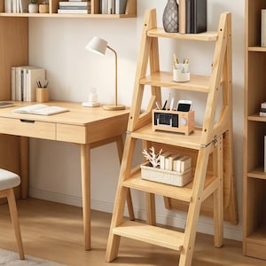 Può includere: Una scala a pioli in legno chiaro con quattro ripiani, utilizzata come libreria. I ripiani contengono libri, oggetti decorativi e una piccola radio di legno. Sulla sinistra c'è una scrivania con una lampada e un libro aperto.