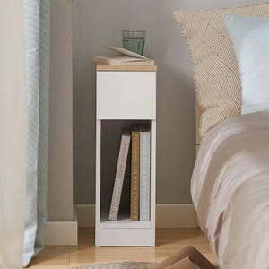 May include: A white bedside table with a light wood top and a small drawer. The table has an open shelf with two books. A glass of water and some papers sit on top. The table is next to a bed.
