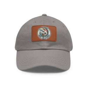 May include: A gray baseball cap with a curved brim. The cap features a brown leather patch with a circular design of a fish jumping out of the water, with the words "HOOK, LINE, & SINKER" in a circular pattern.