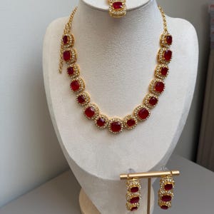 May include: A gold-toned jewellery set featuring a necklace, a pair of earrings, and a maang tikka. Each piece is adorned with square-cut red stones surrounded by small white crystals. The necklace has a chain link design with a lobster claw clasp.