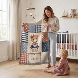 Peut inclure: Une couverture patchwork pour bébé avec un motif d'ours en peluche. La couverture présente des carrés bleus, blancs et marron avec des pois, des rayures et des motifs d'ancres. Le carré central représente un ours en peluche portant une tenue de marin et le nom "Olivia".