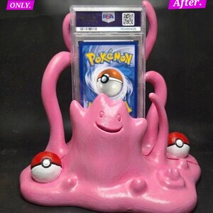 May include: A pink, hand-painted Ditto-themed card holder with two Poké Balls and a graded Pokémon card. The holder has a smiling Ditto face and curved tentacles. The card is in a clear plastic case.
