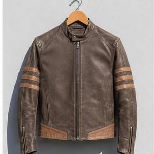 Könnte beinhalten: Eine braune Lederjacke mit Stehkragen und Druckknopfverschluss, durchgehendem Frontreißverschluss und zwei horizontalen beigefarbenen Streifen an jedem Ärmel. Die Jacke verfügt über beigefarbene Ledereinsätze an der Taille und Reißverschlüsse.