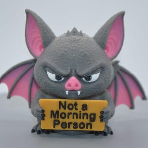 May include: A gray bat figurine with large pointed ears featuring pink inner details, a pink nose, and two white fangs. The bat has a grumpy facial expression and holds a yellow rectangular sign with the black text Not a Morning Person.