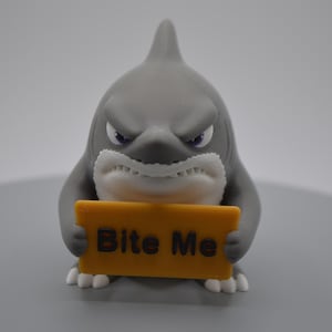 May include: A gray shark figurine with a white underbelly and a stern facial expression. The shark is holding a rectangular yellow sign in its front fins that features the text "Bite Me" printed in dark brown letters on the front surface.