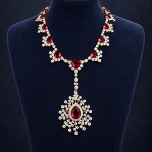 Peut inclure: Un collier doré orné d'une série de pierres rouges en forme de poire entourées de petits cristaux transparents. Le design comprend un pendentif central avec une grande pierre rouge en forme de poire, entourée d'un éclat de cristaux.