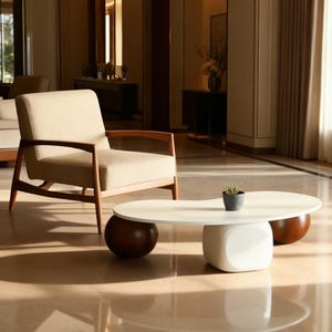 May include: A contemporary living room with a beige armchair and wooden frame. A white, bean-shaped coffee table with dark brown spherical supports and a white cube base holds a small potted plant. The room has a neutral colour palette and polished floor.