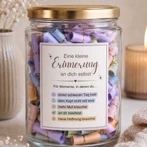 Könnte beinhalten: Ein klares Glas mit goldenem Deckel, gefüllt mit bunten, gerollten Papiernotizen. Ein weißes Etikett mit goldenem Text liest sich "Eine kleine Erinnerung an dich selbst" und listet Gründe auf, die Notizen zu lesen. Die Notizen sind in Rosa, Blau und Gelb gehalten.