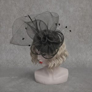 Könnte beinhalten: Ein schwarzer Fascinator mit einer breiten, transparenten Netzbasis, strukturierten Schlaufen und feinen Federakzenten. Das Kopfstück wird auf einem Schaufensterpuppenkopf mit welligem blondem Haar präsentiert und zeigt ein geschichtetes Design.