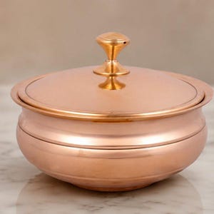 May include: A copper-colored, lidded bowl with a rounded base and a decorative handle on the lid. The bowl has a polished finish, reflecting the light. The design is simple and elegant, suitable for serving or storage.