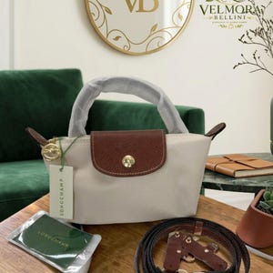 May include: A cream-colored handbag with a brown flap and handle. The bag has a gold clasp and a Longchamp tag. A matching brown strap and accessories are also visible. The bag is on a wooden table.